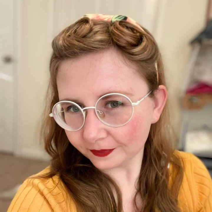 selfie of woman with yellow sweater, white glasses, red lipstick, blond hair in victory rolls.